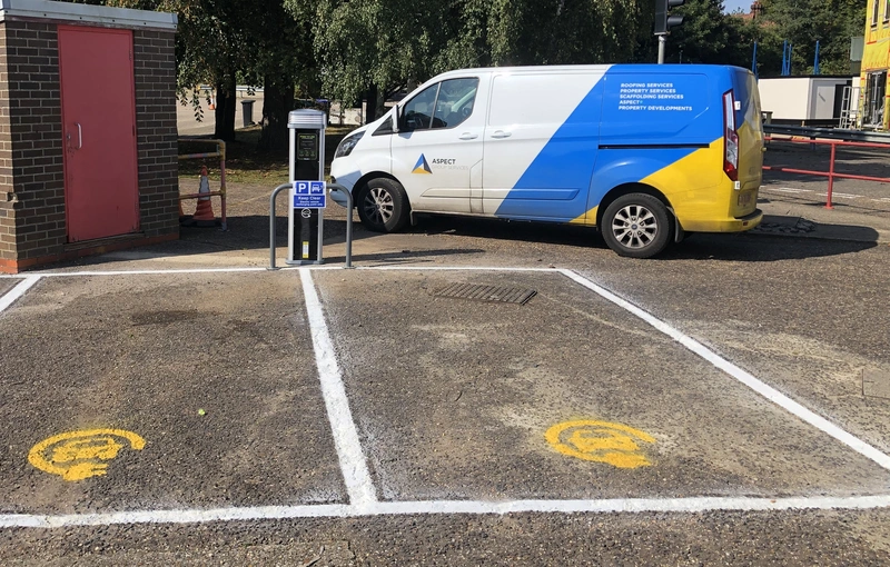 A van with a yellow lower half and blue upper half is parked diagonally, next to two electric vehicle charging spaces in a lot.