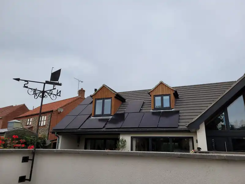 Solar panels installed on the roof of a residential house with dormer windows under a cloudy sky.