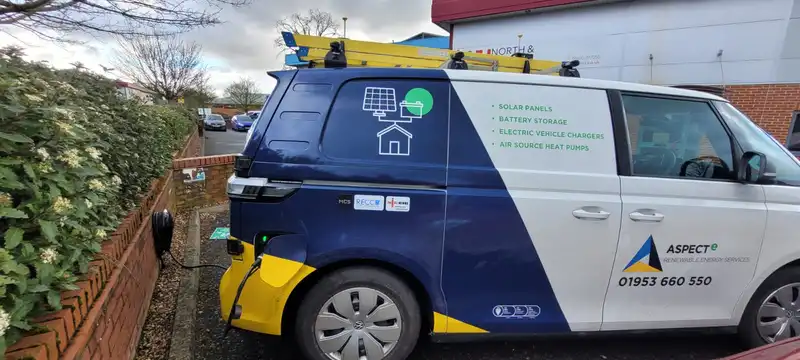 A parked commercial van with solar panels, battery storage, and electric vehicle charger advertisements on the side, equipped with ladders on top.