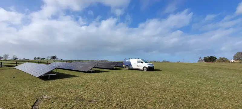 A white electric van parked next to large solar panels in a spacious green field under a partly cloudy sky.