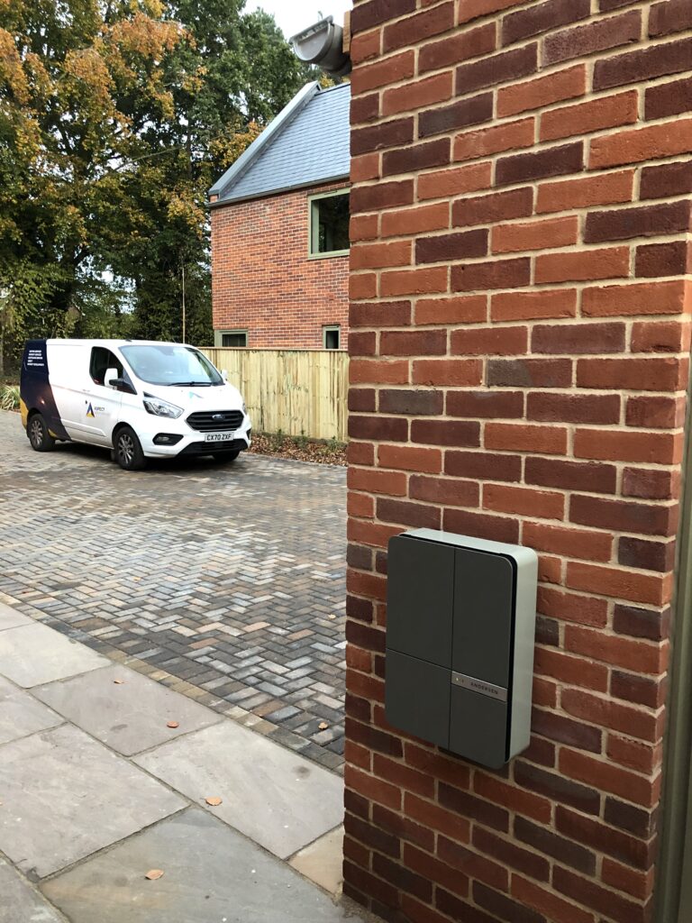 A white van is parked on a cobblestone driveway next to a brick building with an EV charger mounted on the wall. A house with a gray roof is visible in the background.