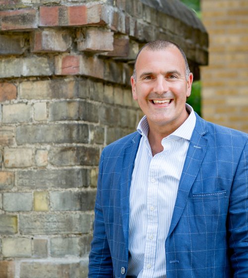 Matt Dillon smiling in a blue blazer and striped shirt standing in front of a brick wall.