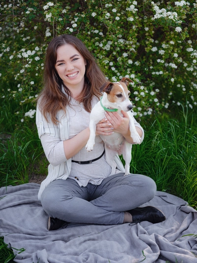 Sarah Johnson smiling, sitting on a blanket outdoors, holding a jack russell terrier, with a backdrop of blooming bushes.