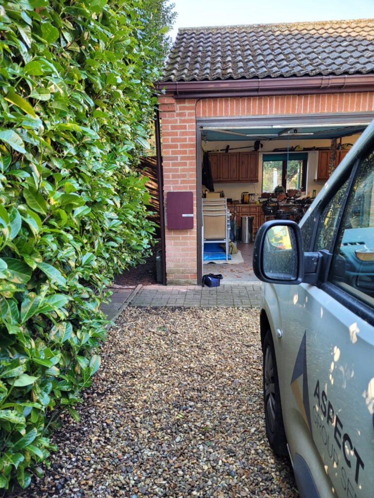 A driveway with a gravel surface leading to a garage partially opened showing stored items, flanked by green foliage and a parked car marked "aspect.