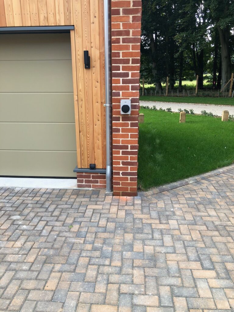 A driveway paved with bricks leads to a garage with a wooden and brick exterior. A black EV charger is mounted on a brick pillar next to the garage. A green lawn with a gravel path is in the background.