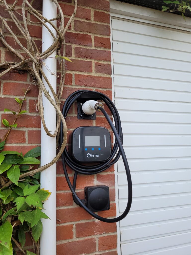 Electric car charger mounted on a brick wall beside a garage door, with the cable neatly coiled around it and a vine climbing beside it.