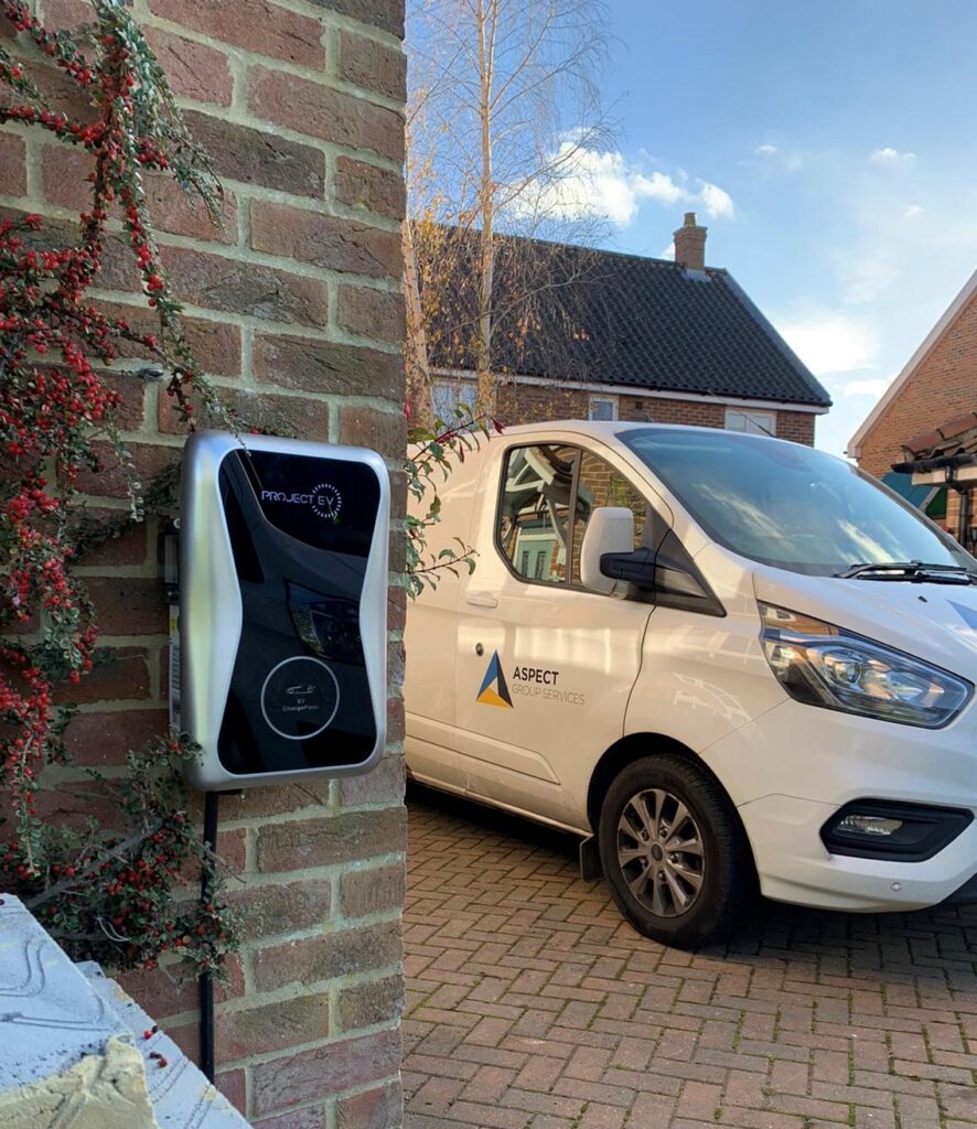 An electric vehicle charging station mounted on a brick wall beside a white van labeled "aspect facades" parked in a driveway, with ivy and a clear blue sky in the background.