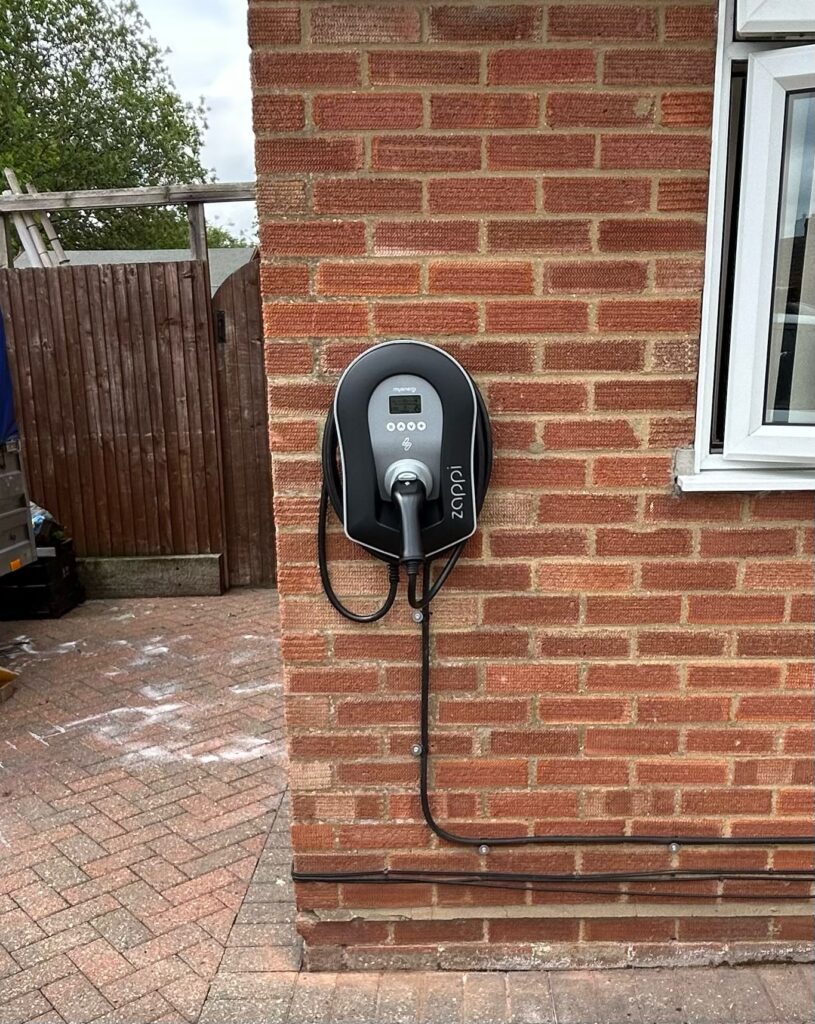 Electric car charging station mounted on a brick wall beside a house window, with a coiled charging cable.