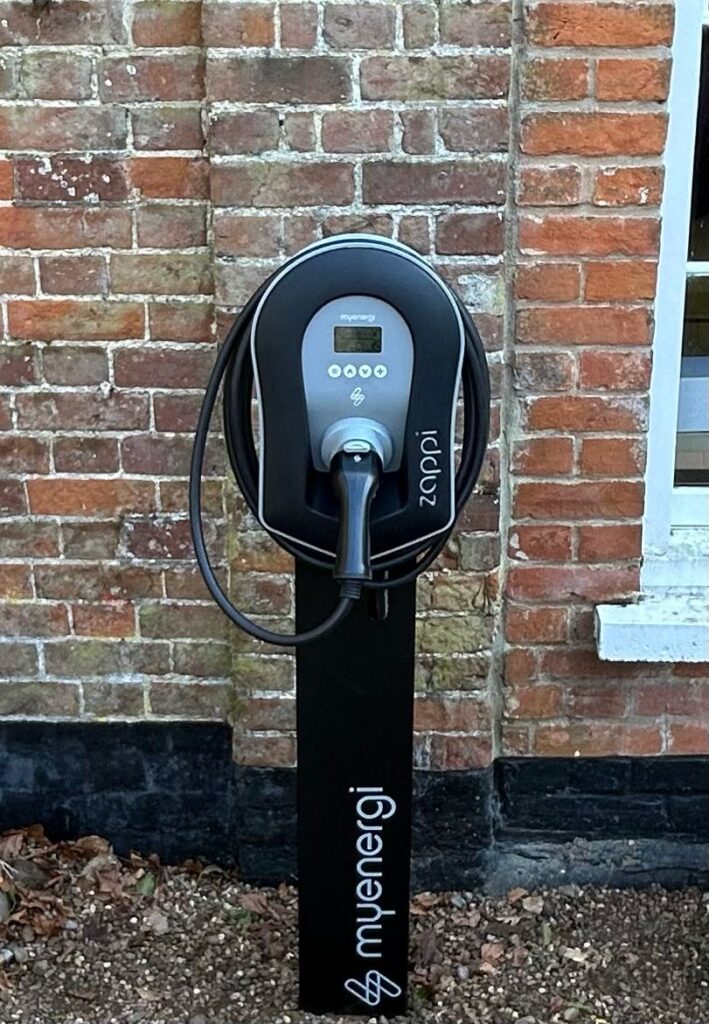 An electric vehicle charging station by murengi, featuring a black stand with a digital display and a connected charging cable, installed beside a brick wall.